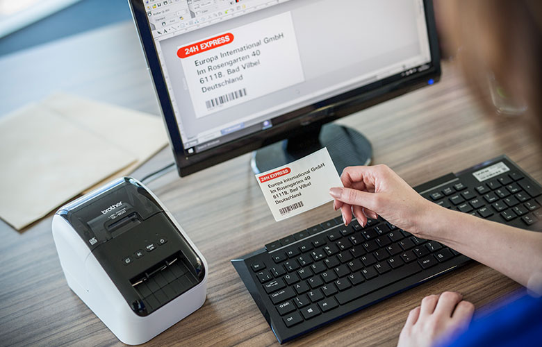 Brother QL tiskárna vedle monitoru s osobou držící štítek s adresou v kanceláři