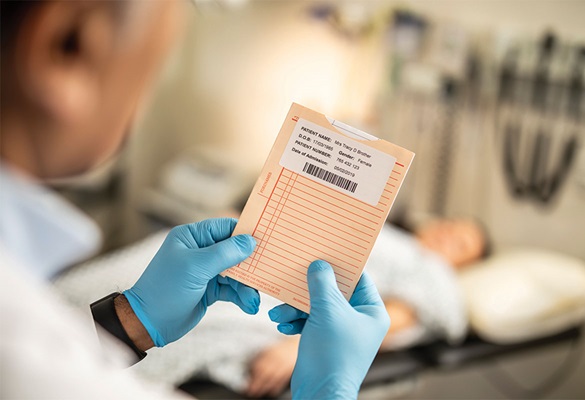 Doktor s modrými rukavicemi drží soubor pacientky s jejími detaily na štítku, pacientka leží na nemocničním lůžku